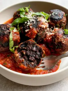 Close up handheld photo of five vegan black bean meatballs in a white bowl showing charred edges and flecks of cilantro.